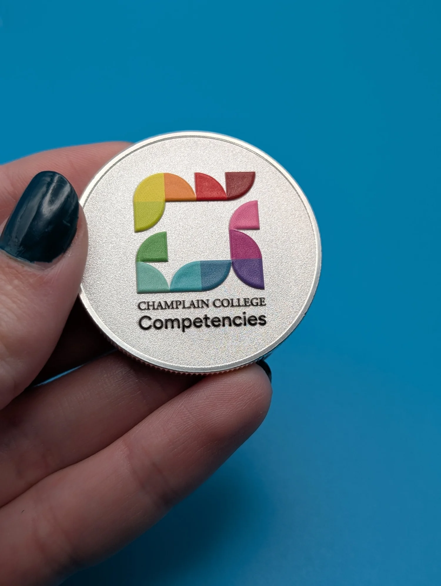 A UV-printed metal coin with a full-color Champlain College Competencies mark, held in hand.